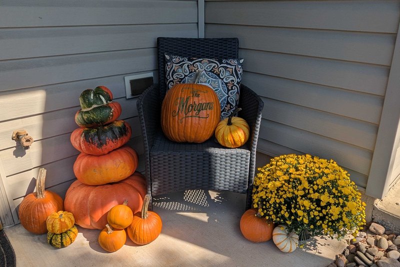 The Signature Porch in all orange on a real porch corner: a stack of orange and green heirloom pumpkins, a wicker chair holding a pumpkin engraved The Morgans, a yellow mum and small pumpkins around them